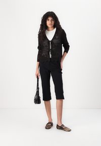 Melns atvērtas adīšanas kardigans ar sasienamu aizdari, virs balta topa. Valkāts ar melnām īsām biksēm un leoparda raksta balerīnām, turēt mazu melnu somiņu.