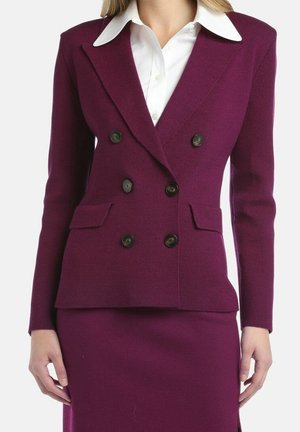 Kvinde iført en tætsiddende blommefarvet dobbeltradet blazer med sorte knapper over en hvid skjorte med krave og matchende blommefarvet nederdel.