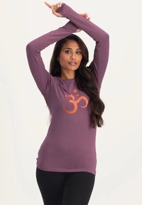 Langärmliges, lila Oberteil aus strukturiertem Stoff mit orangefarbenem "Om"-Symbol-Druck in der Mitte, kombiniert mit schwarzen Leggings.