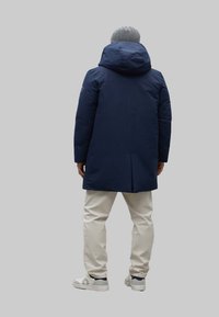 Parka aislante de color azul marino con capucha, que presenta una apertura trasera, tejido texturizado y se complementa con pantalones de color crema y zapatillas blancas.
