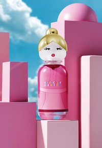 Pink United Colors of Benetton parfume flaske formet som en stiliseret kvinde med gyldent hår, røde læber, på pink geometriske blokke mod blå himmel.