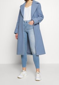 Ljusblå kappa med spetsig krage, oversized ärmar och öppen framsida, bärs över en vit topp och ljusblå tajta jeans. Vita sneakers.