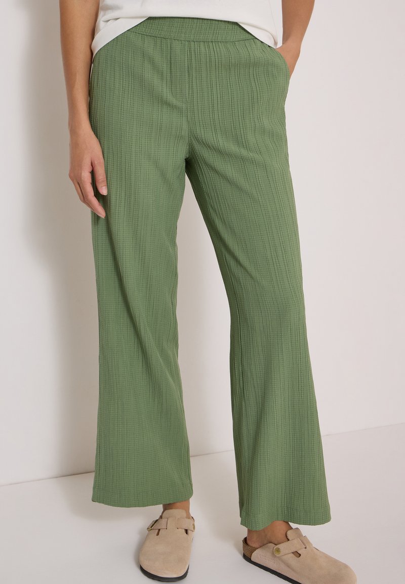 Femme portant un pantalon ample vert clair à rayures verticales texturées, associé à des chaussures beige sans lacets, debout devant un fond uni.