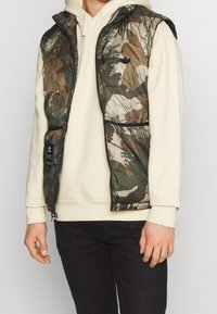 Kamuflażowa kamizelka z zamek błyskawicznym z przodu, lekkim wypełnieniem i kapturem. Charakteryzuje się mieszanką zieleni i brązów, ozdobiona wzorami gałęzi.