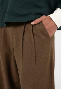 Bruine wollen broek met zijzakken, voorzien van plooien en een gestructureerde afwerking, gecombineerd met een donkergroen shirt. Hand rustend op de broek.