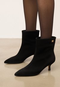 Botine negre din suede, cu vârfuri ascuțite, toc de înălțime medie și un design cu mansetă pliată. Detaliu cu logo auriu pe partea exterioară.