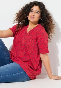 Maglietta rossa con scollatura a V e maniche corte, caratterizzata da un design a motivi neri. Abbinata a jeans blu, si trova su uno sfondo bianco.