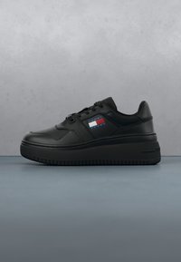 Basket en cuir noir avec semelle épaisse et patch logo Tommy Jeans sur le côté, présenté sur un fond gris uni.