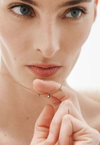 Collar de oro delicado con tres pequeñas gemas brillantes. La persona sostiene el collar cerca de su rostro, con el cabello corto y un fondo neutro.