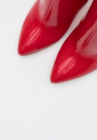 Błyszczące czerwone botki z ostrym noskiem i centralnym szwem na białym tle, podkreślające błyszczącą teksturę i smukły design.