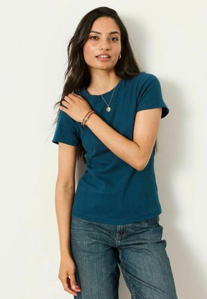 Giovane donna con lunghi capelli scuri indossa una maglietta verde acqua a maniche corte, jeans blu, una collana d'oro e braccialetti di perline, posando davanti a una parete semplice.