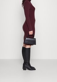 Burgundská žebrovaná šaty s dlouhými rukávy, doplněné černými kozačkami po kolena a strukturovanou černou kabelkou s horním držadlem.