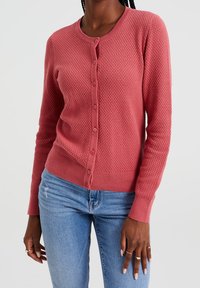 Růžový cardigan s texturovanou látkou, kulatým výstřihem a zapínáním na knoflíky vpředu. Dlouhé rukávy a žebrované manžety dodávají detail.