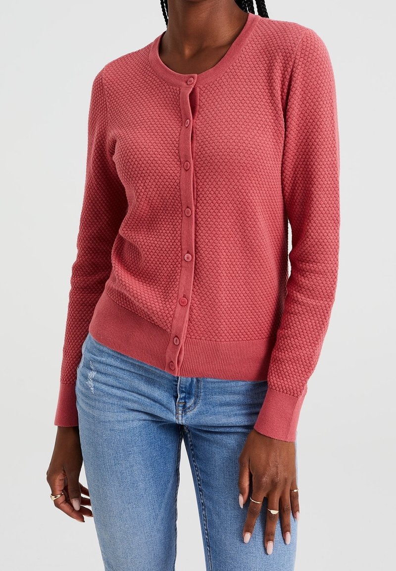 Růžový cardigan s texturovanou látkou, kulatým výstřihem a zapínáním na knoflíky vpředu. Dlouhé rukávy a žebrované manžety dodávají detail.