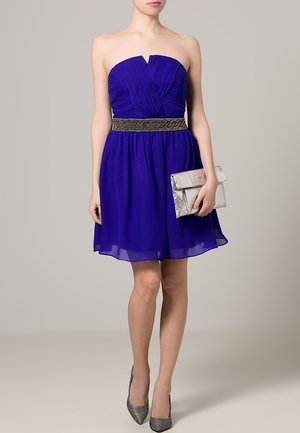 Robe violette sans bretelles avec des plis, ceinture argentée ornée, et jupe fluide. Associée à une pochette argentée et des talons gris.