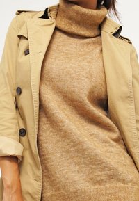 Manteau beige à gros boutons sur un pull encolure montant tan. Les textures sont lisses et douces, avec une silhouette ajustée.