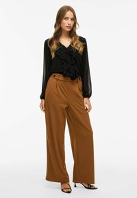 Blusa nera con volant e maniche lunghe trasparenti, abbinata a pantaloni larghi a vita alta marroni con due bottoni frontali e una texture liscia.