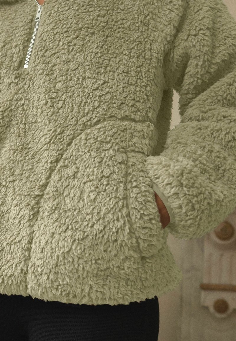 Personne portant une veste zippée duveteuse vert clair avec les mains dans les poches, montrant la texture et le détail de la fermeture éclair avant.