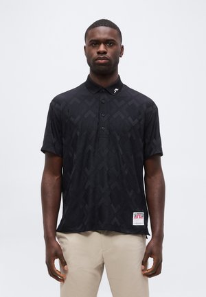 Homme debout portant un polo noir à motifs avec un logo sur le col et un pantalon beige sur un fond clair uni.