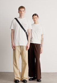 Kaksi henkilöä seisoo vierekkäin, pukeutuneina valkoisiin t-paitoihin, joissa on graafisia kuvioita, yhdistettynä väljiin beigeihin ja tummanruskeisiin housuihin.