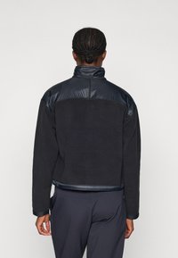 Svart fleecejakke med en blank marineblå øverdel og krage. Har en tettsittende design og glatt tekstur, med en kontrasterende nedre del.