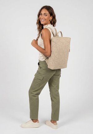 Kvinde iført beige ærmeløs top, olivengrønne cargobukser og hvide sneakers, bærende en beige rygsæk med geometrisk mønster, smilende og kigger tilbage.