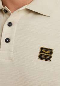 Cremefarvet polo-shirt med tekstureret mønster, der har tre mørke knapper og et PME Legend-brandede mærke på brystet.