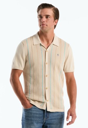 Man in een beige kortebovenknopen-overhemd met verticale strepen en een blauwe spijkerbroek, staand met één hand in de zak, kijkend naar links.