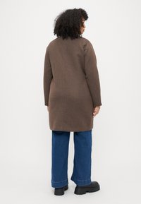 Casaco castanho em mistura de lã, com corte reto, mangas longas e uma abertura nas costas, combinado com calças de ganga azul de perna larga e sapatos de plataforma pretos.