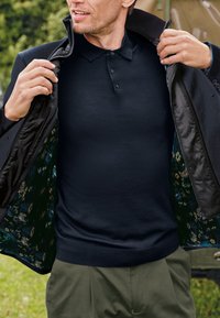 Granatowa koszulka polo pod czarną kurtką, z kwiecistą podszewką. Kurtka ma gładką fakturę i zamek błyskawiczny.