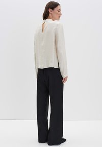 Donna che indossa una blusa a maniche lunghe testurizzata di un bianco sporco con un bottone a occhiello sul retro, abbinata a pantaloni neri a gamba larga, in piedi di profilo.
