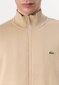 Beige glidelåsjakke med høy krage, laget av mykt materiale. Har et lite brodert grønt krokodillelogo på brystet.