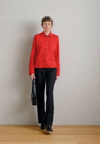 Κόκκινο πουκάμισο με μακριά μανίκια, σε συνδυασμό με μαύρο παντελόνι και μαύρα παπούτσια. Κρατάει μια μαύρη τσάντα, στέκοντας μπροστά σε έναν απλό φόντο.