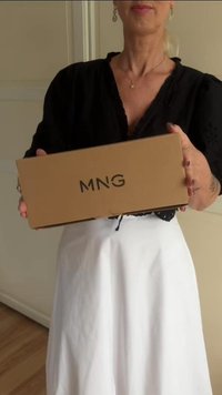 Kobieta w czarnej bluzce i białej spódnicy trzyma brązowy karton z napisem "MNG" w kierunku kamery w pomieszczeniu.