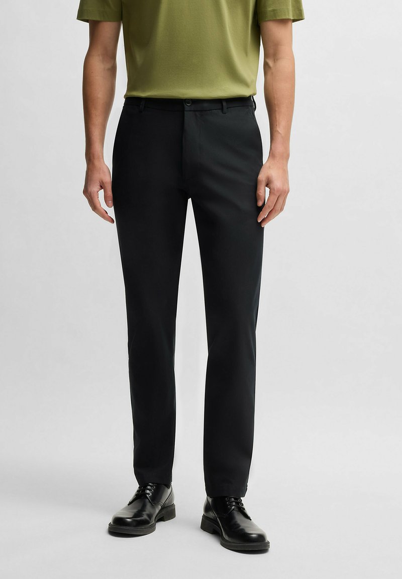 Black trousers with a slim fit, featuring a smooth texture, a front zipper, and button closure. Worn with a green short-sleeve shirt.