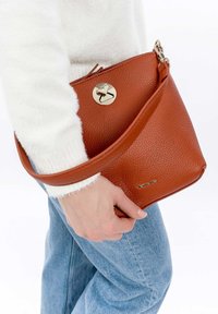 Bolso de mano de cuero marrón con un acabado texturizado, que presenta un cierre circular dorado y una correa para el hombro, sostenido en la mano de una persona.