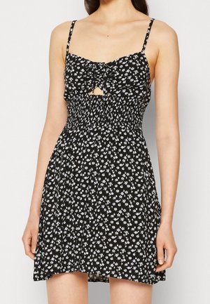 Zwarte mini-jurk met wit bloemenpatroon, dunne bandjes, gerimpelde buste met kleine voorsnede en elastische geplooide taille, gedragen door een vrouw.