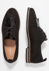 Chaussures à lacets en daim noir avec un accent en cuir verni noir brillant et un design unique à franges sur le bout. Semelle plate avec des détails couleur tan.