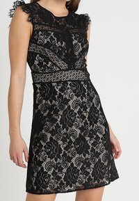Robe noire en dentelle avec motifs floraux, design sans manches, épaules à volants et taille cintrée. Présente une texture semi-transparente subtile.