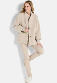 Beige textuur jas met een brede kraag, twee knopen en zijzakken, gecombineerd met lichtbeige broek en witte sneakers.