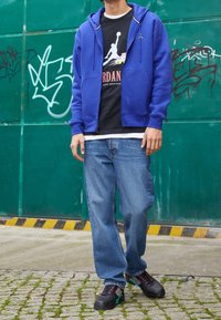 Sudadera con capucha azul con cremallera, camiseta negra con logo, vaqueros azul claro y zapatillas negras con detalles en rosa y turquesa. El suelo es de adoquines.