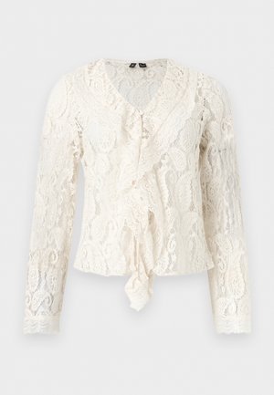 Blouse blanche à manches longues en dentelle avec décolleté en V, détail à volants sur le devant et motifs floraux complexes sur l'ensemble.