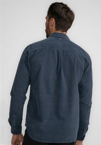 Donkerblauwe overhemd met lange mouwen van een zachte stof, met knopen aan de manchetten en een rugstuk detail. Heeft een subtiele merklabel nabij de hals.