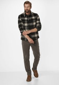 Mann in einer schwarz-weißen karierten Jacke, grauem Hemd, grauen Hosen und braunen Schuhen, der den Ärmel vor einem schlichten weißen Hintergrund anpasst.