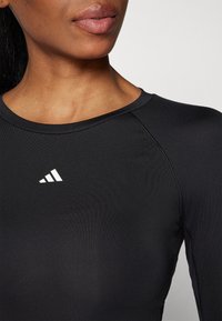 Top de sport noir avec un col rond, un design ajusté et un logo blanc sur le côté gauche. Le tissu semble lisse et extensible.