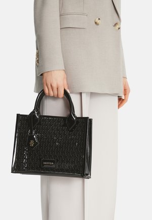 Žena držící černou lesklou kabelku s krátkými držadly, oblečená v béžovém saku a bílých kalhotách na bílém pozadí.