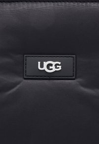 Czarna torba typu tote z nylonu o fakturowanej powierzchni oraz prostokątna naszywka z logo UGG w kolorze białym. Charakteryzuje się prostym, funkcjonalnym designem z minimalnymi akcentami.