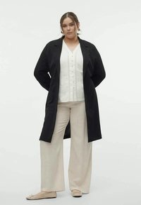 Casaco longo negro sobre uma blusa de malha branca com botões, combinado com calças largas beiges e sapatos planos de tom claro; manequim de pé contra um fundo simples.