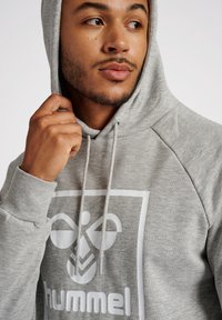 Grijze hoodie met een trekkoord, voorzien van een groot wit logo aan de voorkant. Het materiaal lijkt zacht, met gestructureerde mouwen en een relaxte pasvorm.