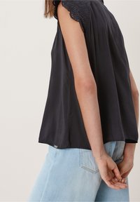 Blusa nera senza maniche con dettagli in pizzo sulle spalle, tessuto morbido e vestibilità rilassata, abbinata a jeans azzurri chiari.
